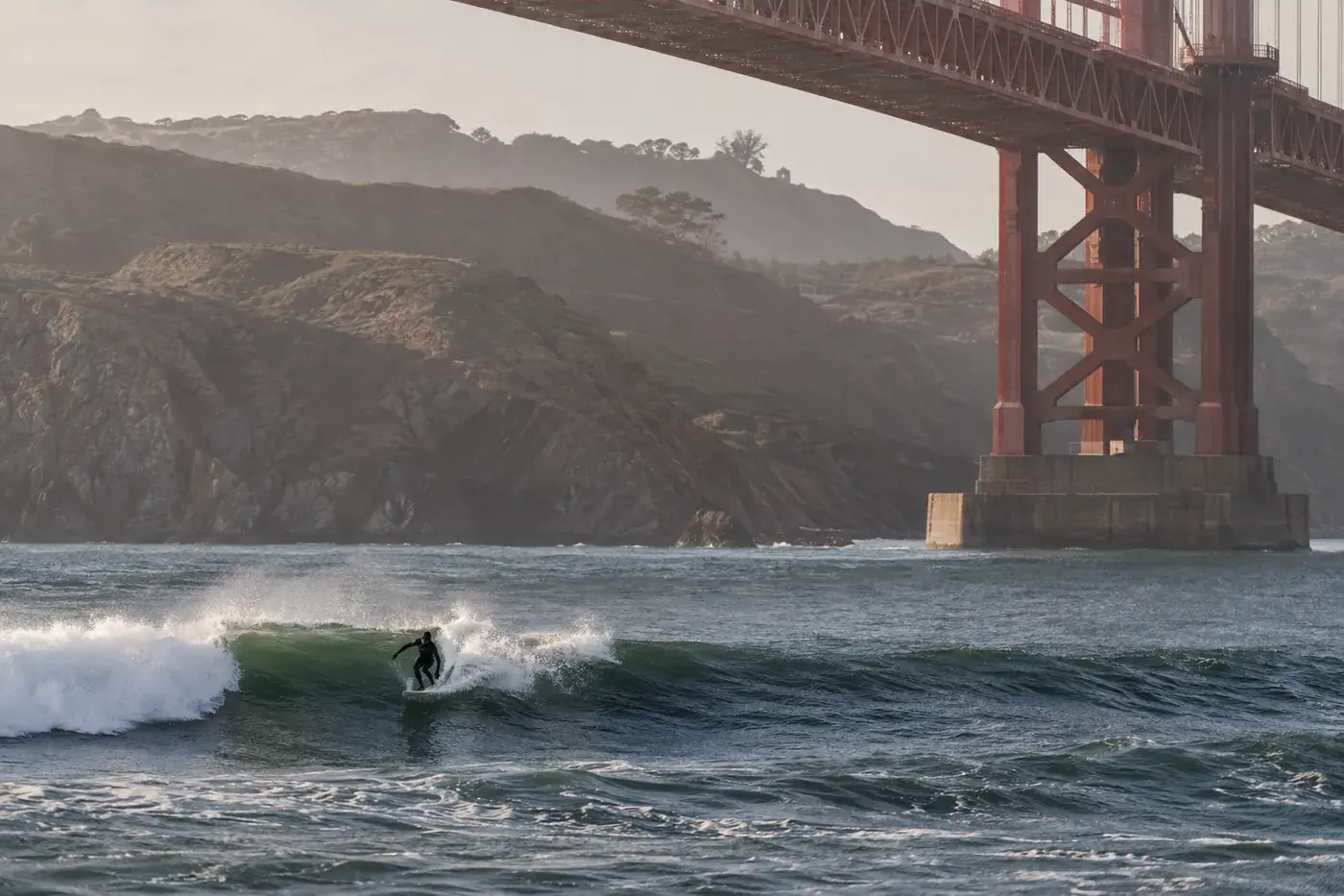 A distant surfer riding a small wave beneath cropped bridge steel.